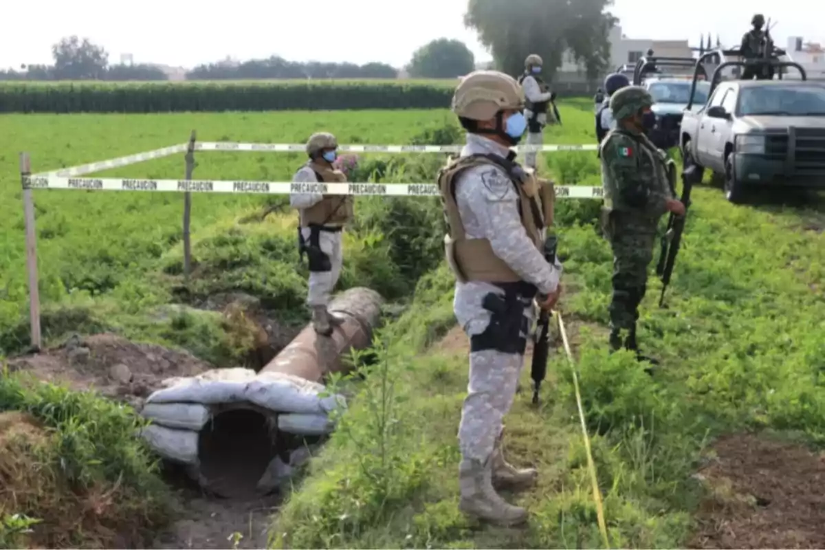 Soldiers with tactical gear guard an area cordoned off with caution tape in a green field.