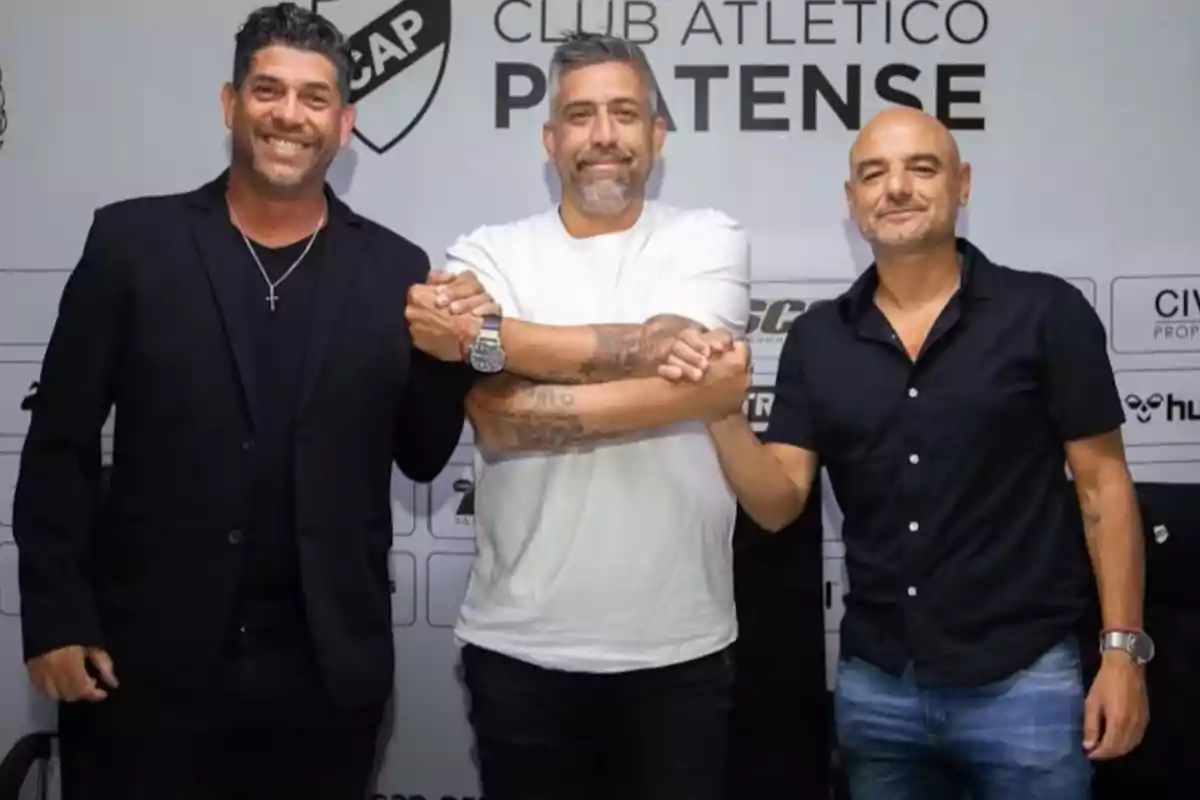 Tres hombres posan sonrientes frente a un fondo con el logo y nombre del Club Atlético Platense mientras se toman de las manos en señal de unión Tres hombres posan sonrientes frente a un fondo con el logo y nombre del Club Atlético Platense mientras se toman de las manos en señal de unión