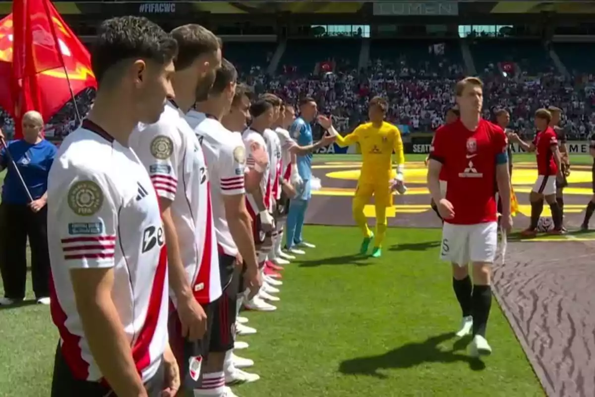 Players from two soccer teams greet each other on the field before starting a match, with spectators in the stands and a red flag in the background.