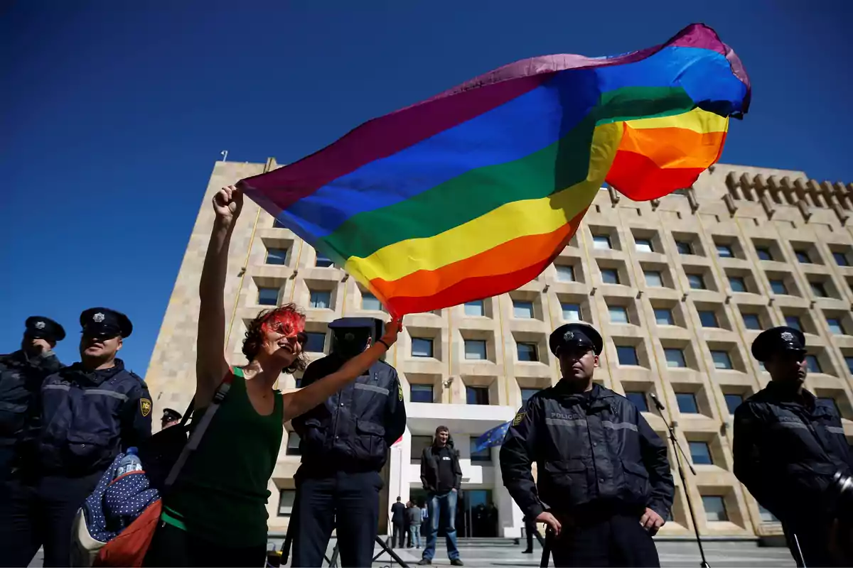 Persona sosteniendo una bandera del arcoíris frente a un edificio con varios policías alrededor.