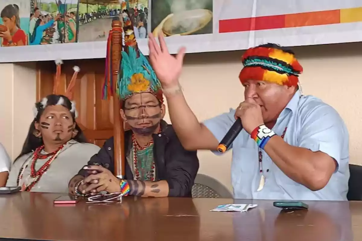 Three people in traditional indigenous clothing and adornments are sitting at a table, one of them holding a microphone while speaking passionately.