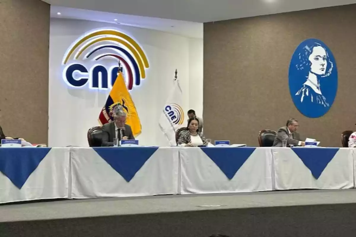 A group of people is sitting at a long table with white and blue tablecloths, behind them is a large logo on the wall and an Ecuadorian flag, as well as a portrait in a blue oval.