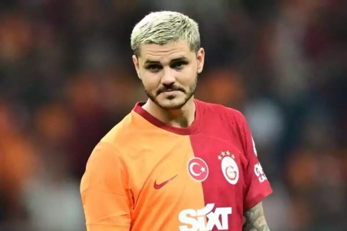 Un jugador de fútbol con cabello rubio y barba lleva una camiseta roja y naranja en un estadio.