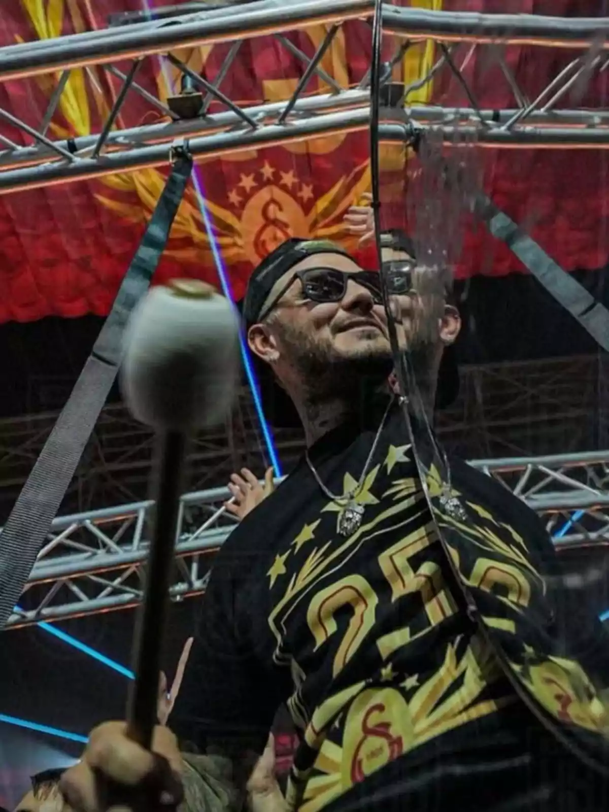 Un hombre con gafas de sol y gorra sostiene un micrófono en un escenario con luces y una estructura metálica, mientras una bandera roja con un emblema amarillo cuelga en el fondo. Un hombre con gafas de sol y gorra sostiene un micrófono en un escenario con luces y una estructura metálica, mientras una bandera roja con un emblema amarillo cuelga en el fondo.