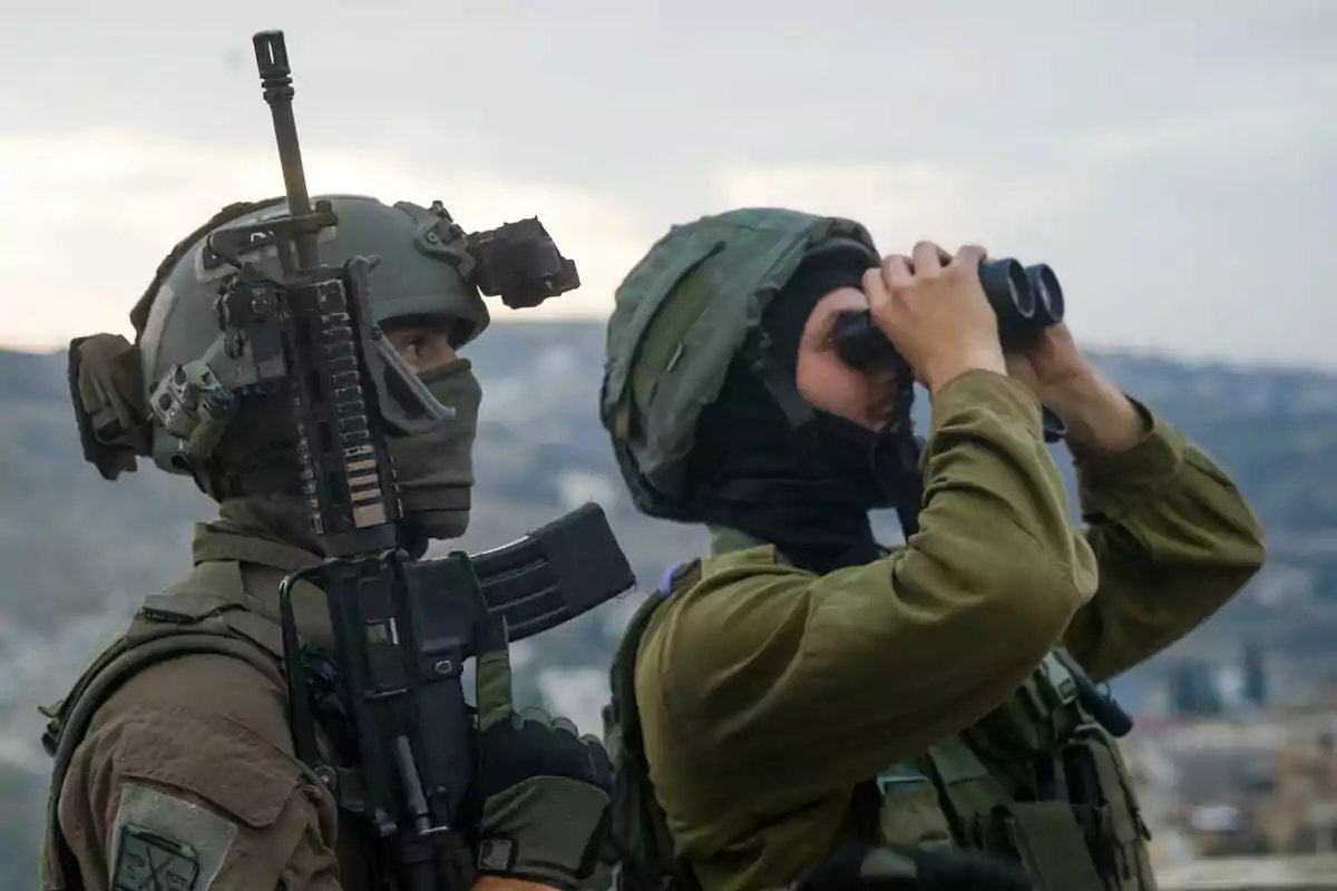 Dos soldados con equipo militar observan el paisaje; uno sostiene un rifle y el otro usa binoculares.