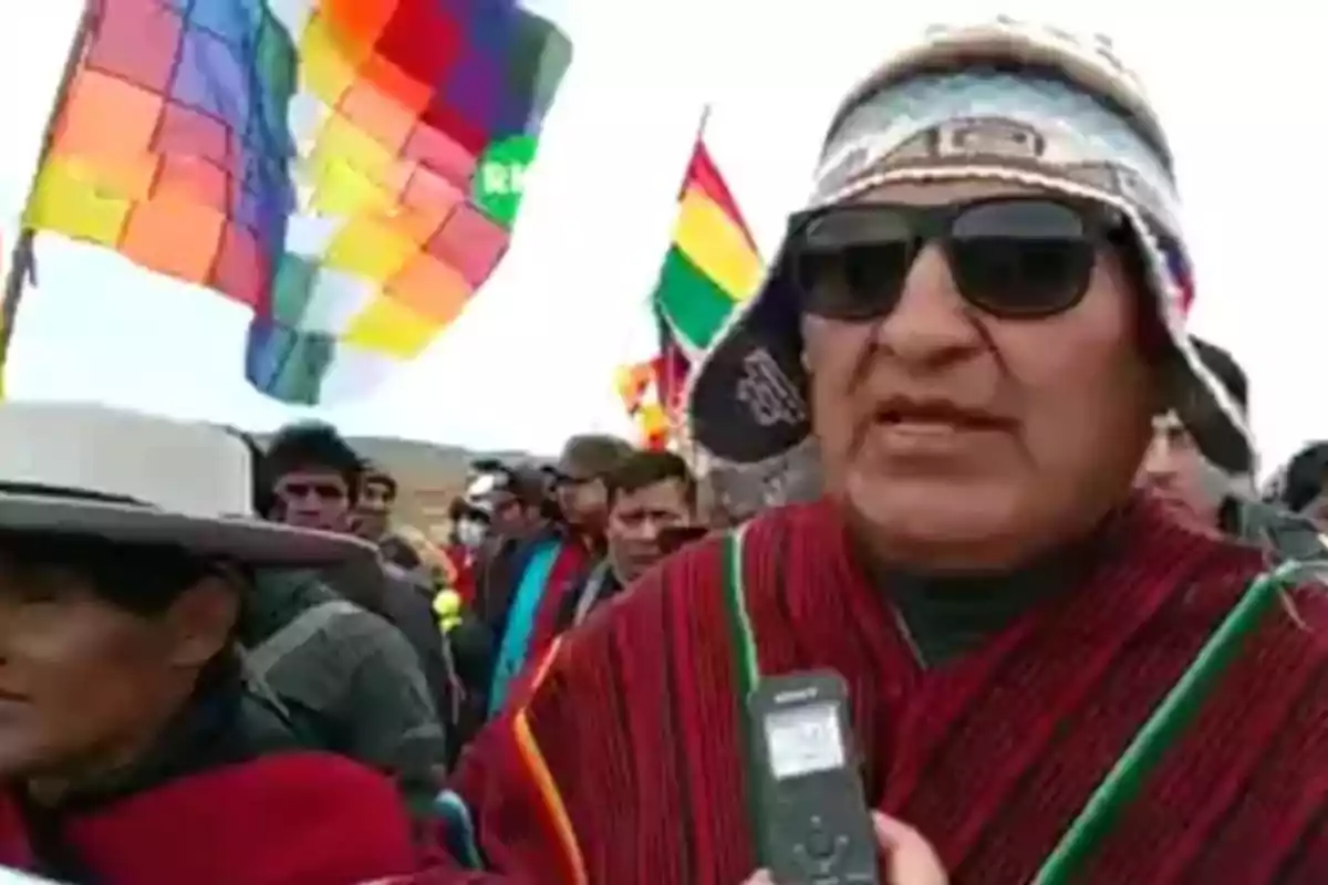 Un grupo de personas, algunas con vestimenta tradicional y banderas coloridas, participa en un evento al aire libre.