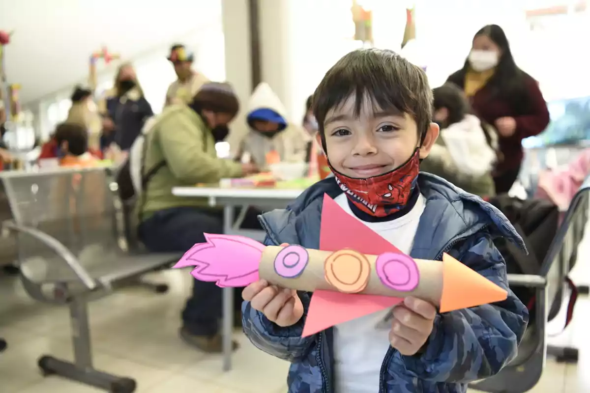 Niño sonriente sostiene una manualidad de cohete hecha con materiales reciclados en un salón donde otras personas también participan en actividades creativas.