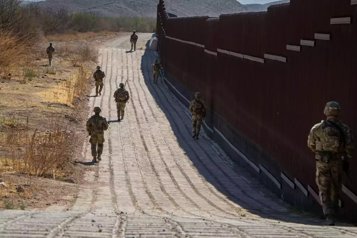 Several soldiers patrol on foot next to a border fence in a desert area.