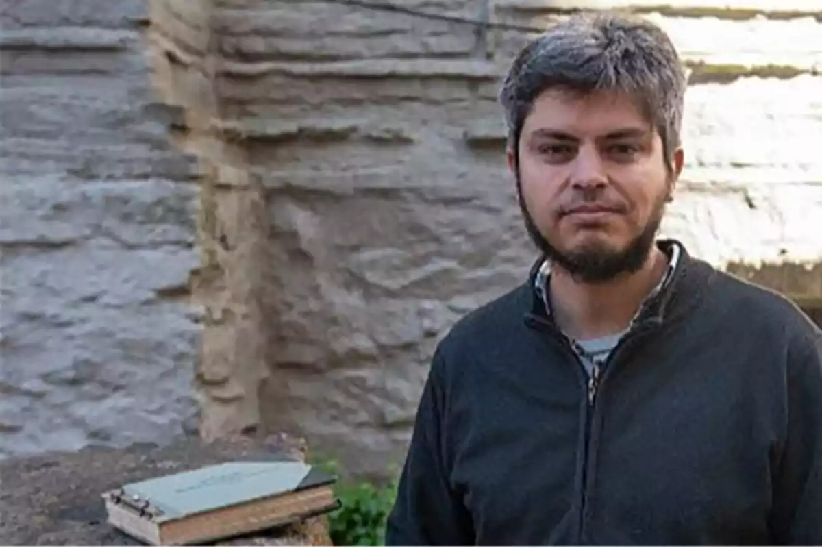 A man standing next to a stone wall with a book on a nearby surface.