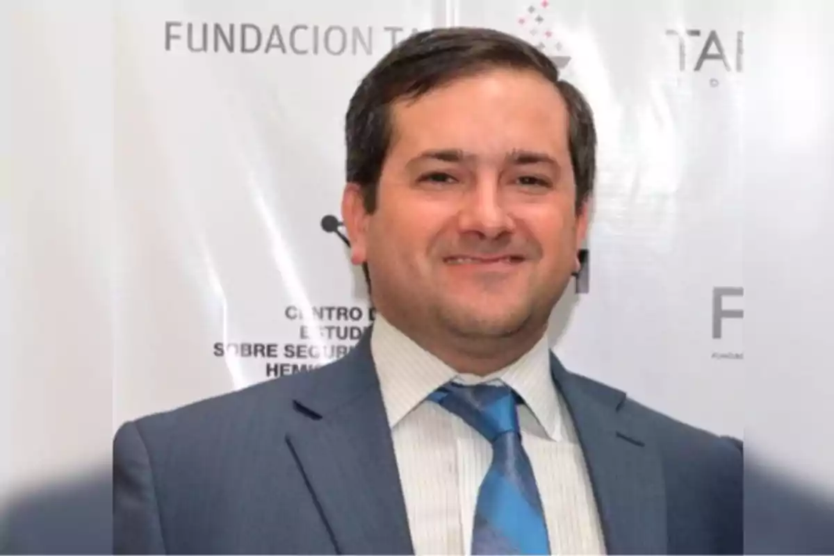A man in a suit and tie smiles in front of a background with foundation logos. A man in a suit and tie smiles in front of a background with foundation logos.