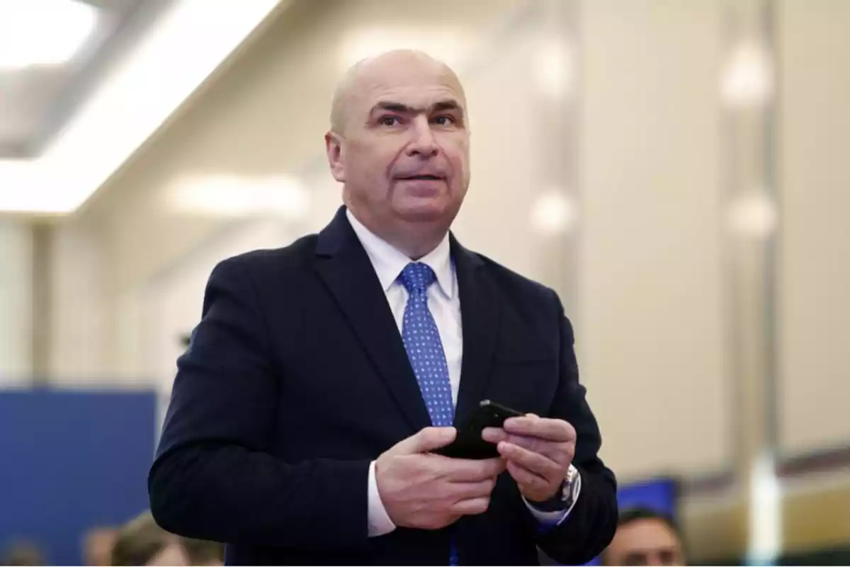 A man in a dark suit and blue tie holds a mobile phone in a well-lit indoor setting. A man in a dark suit and blue tie holds a mobile phone in a well-lit indoor setting.