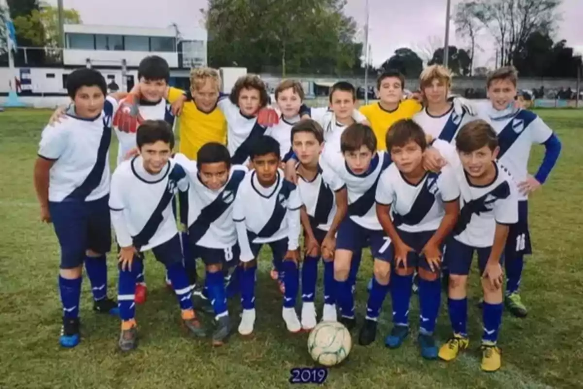 Un equipo de fútbol infantil posa junto a una pelota en una cancha de césped, todos llevan uniforme blanco con una franja azul y medias azules, al fondo se ve una tribuna y árboles Un equipo de fútbol infantil posa junto a una pelota en una cancha de césped, todos llevan uniforme blanco con una franja azul y medias azules, al fondo se ve una tribuna y árboles