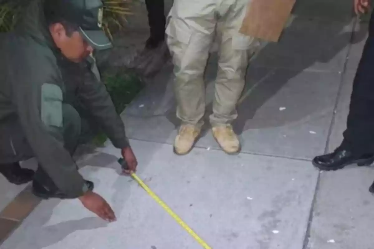 Un policía mide una distancia en la acera con una cinta métrica mientras otras personas observan. Un policía mide una distancia en la acera con una cinta métrica mientras otras personas observan.