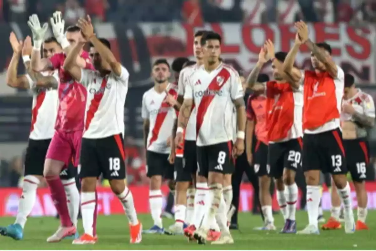 Jugadores de fútbol con uniforme blanco y rojo aplaudiendo en un estadio.