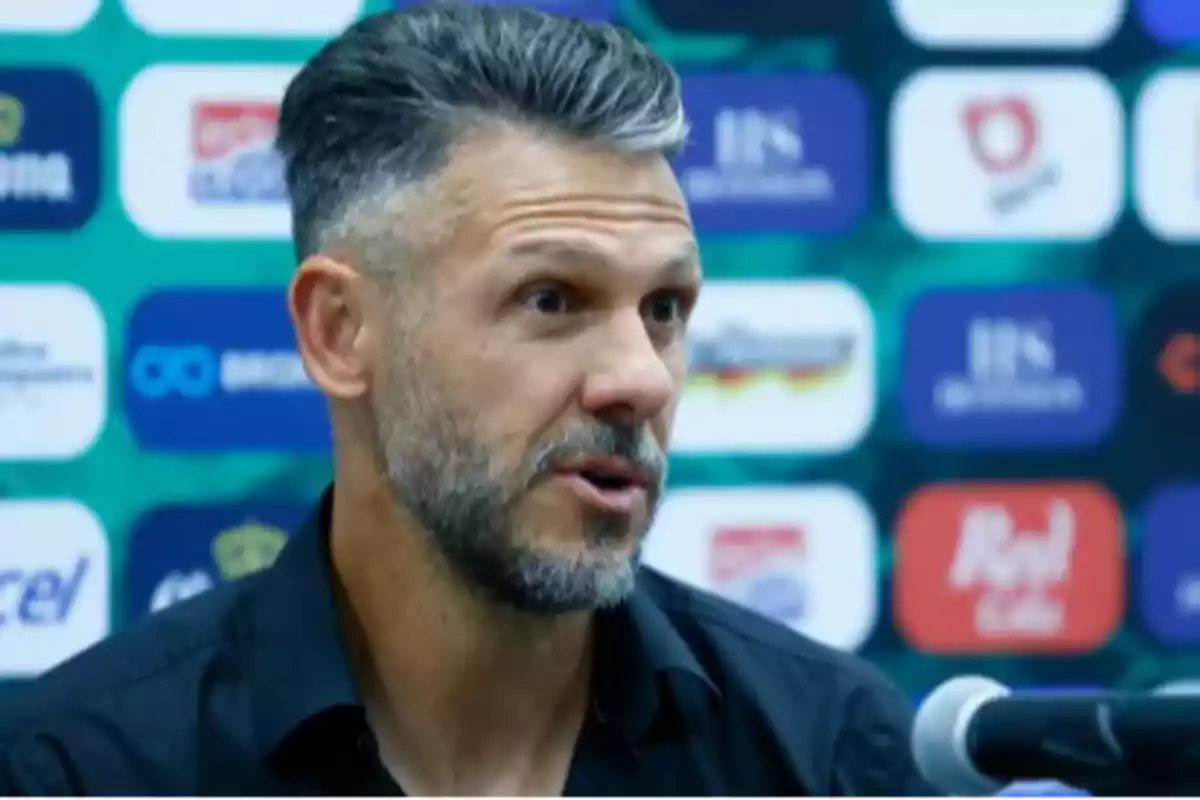Un hombre con barba y cabello corto habla frente a un micrófono con un fondo de logotipos. Un hombre con barba y cabello corto habla frente a un micrófono con un fondo de logotipos.