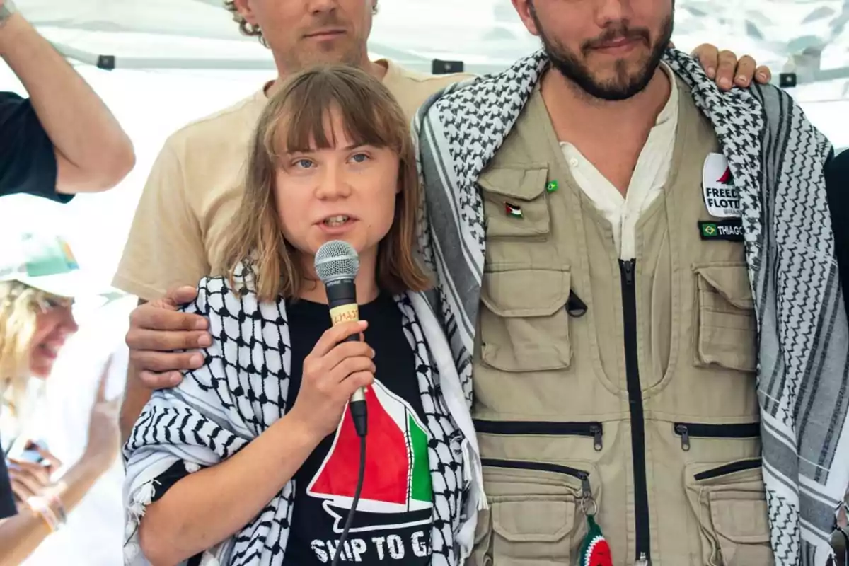 Young woman with a microphone and a Palestinian scarf surrounded by people at an outdoor event