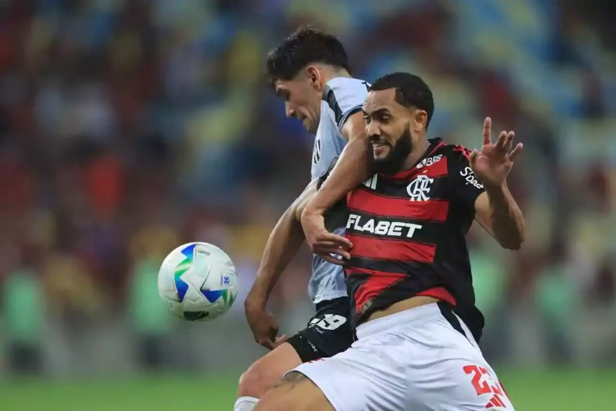 Two players compete for the ball during a match, one wearing a black and red uniform and the other wearing a gray uniform. Two players compete for the ball during a match, one wearing a black and red uniform and the other wearing a gray uniform.