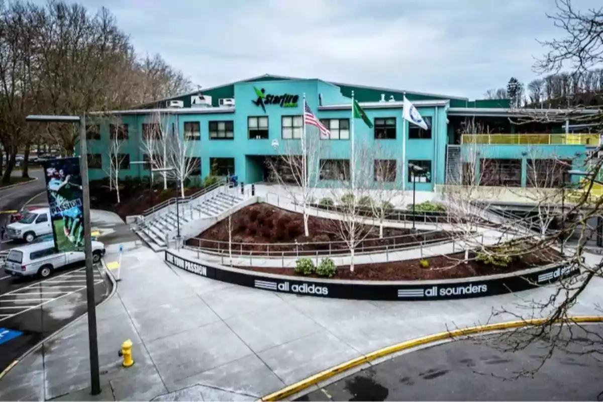 Edificio de color verde agua con letrero de Starfire Sports al frente, tres banderas ondeando y árboles sin hojas en la entrada, con rampas y escaleras rodeadas de anuncios de Adidas y Sounders Edificio de color verde agua con letrero de Starfire Sports al frente, tres banderas ondeando y árboles sin hojas en la entrada, con rampas y escaleras rodeadas de anuncios de Adidas y Sounders