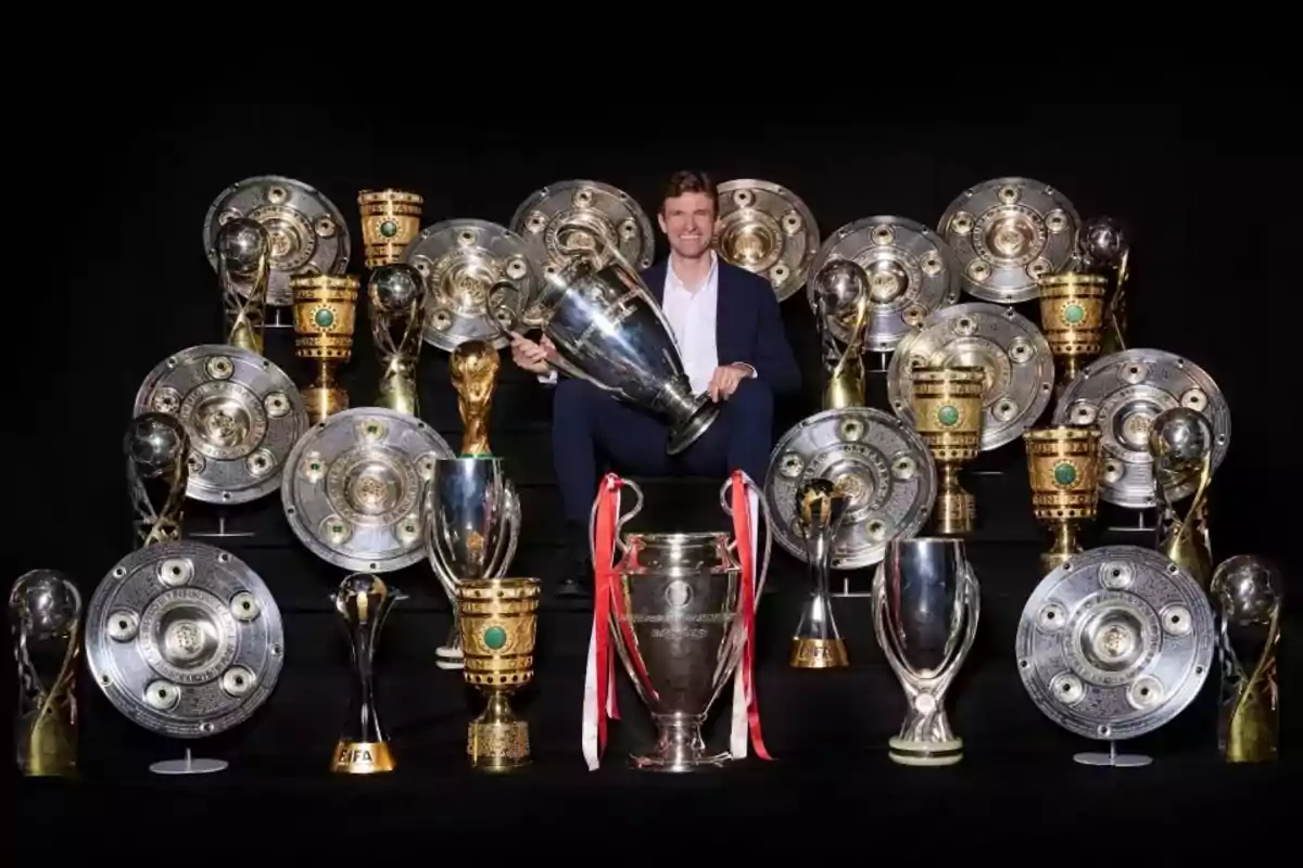 Un hombre con traje sonríe mientras está rodeado de numerosos trofeos de fútbol, incluyendo copas y escudos plateados, todos sobre un fondo negro Un hombre con traje sonríe mientras está rodeado de numerosos trofeos de fútbol, incluyendo copas y escudos plateados, todos sobre un fondo negro