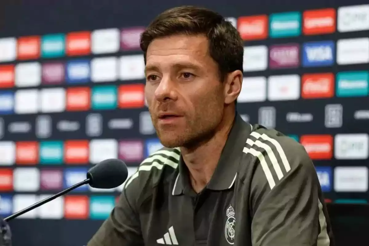 Man wearing a Real Madrid sports jersey speaking at a press conference in front of a microphone and a sponsor backdrop. Man wearing a Real Madrid sports jersey speaking at a press conference in front of a microphone and a sponsor backdrop.