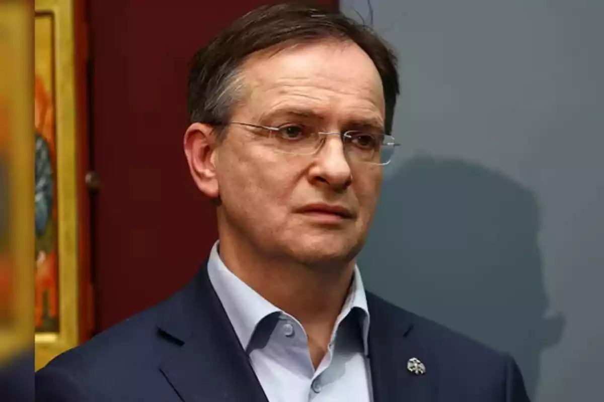 Un hombre con gafas y traje oscuro está de pie frente a un fondo de colores neutros. Un hombre con gafas y traje oscuro está de pie frente a un fondo de colores neutros.