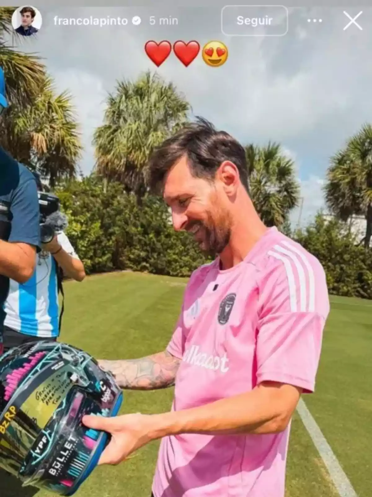 A player in a pink jersey smiles while holding a decorated helmet on a grass field surrounded by palm trees. A player in a pink jersey smiles while holding a decorated helmet on a grass field surrounded by palm trees.