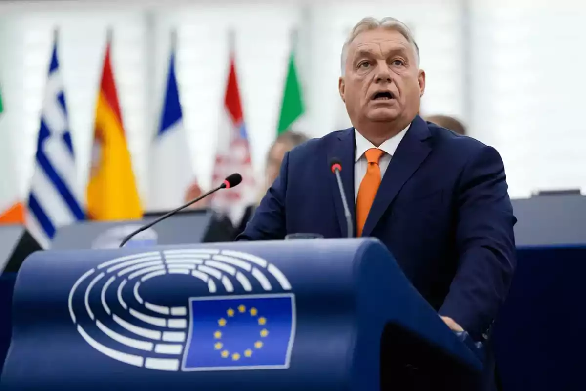 A man in a blue suit and orange tie speaks at a podium with the emblem of the European Union, with several country flags in the background. A man in a blue suit and orange tie speaks at a podium with the emblem of the European Union, with several country flags in the background.