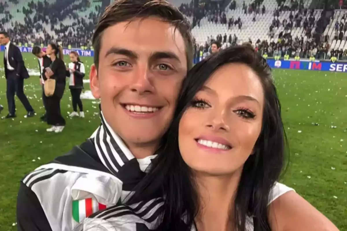 Una pareja sonriente se toma una selfie en un estadio de fútbol después de un partido, con personas y asientos en el fondo.