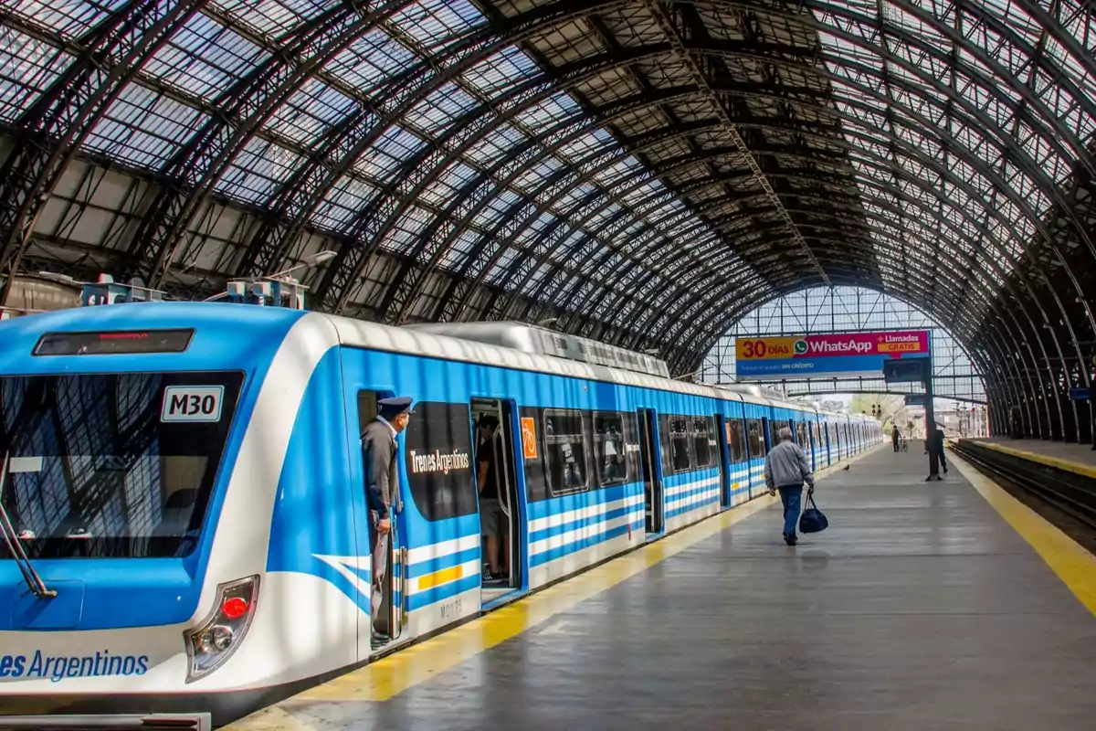 Un tren de Trenes Argentinos en una estación con techo de estructura metálica y personas en el andén.