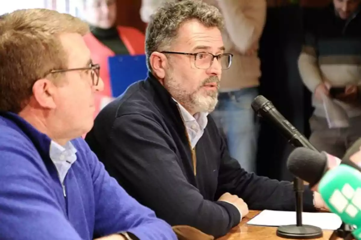 Dos hombres sentados en una mesa durante una conferencia de prensa, uno de ellos hablando frente a un micrófono.