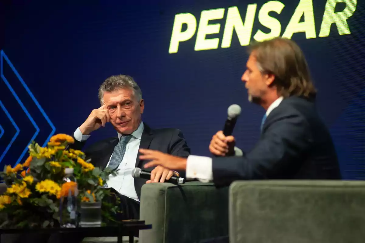 Dos hombres en un escenario participando en una conversación, uno de ellos sostiene un micrófono mientras el otro escucha atentamente, con un fondo que tiene la palabra "PENSAR" iluminada.