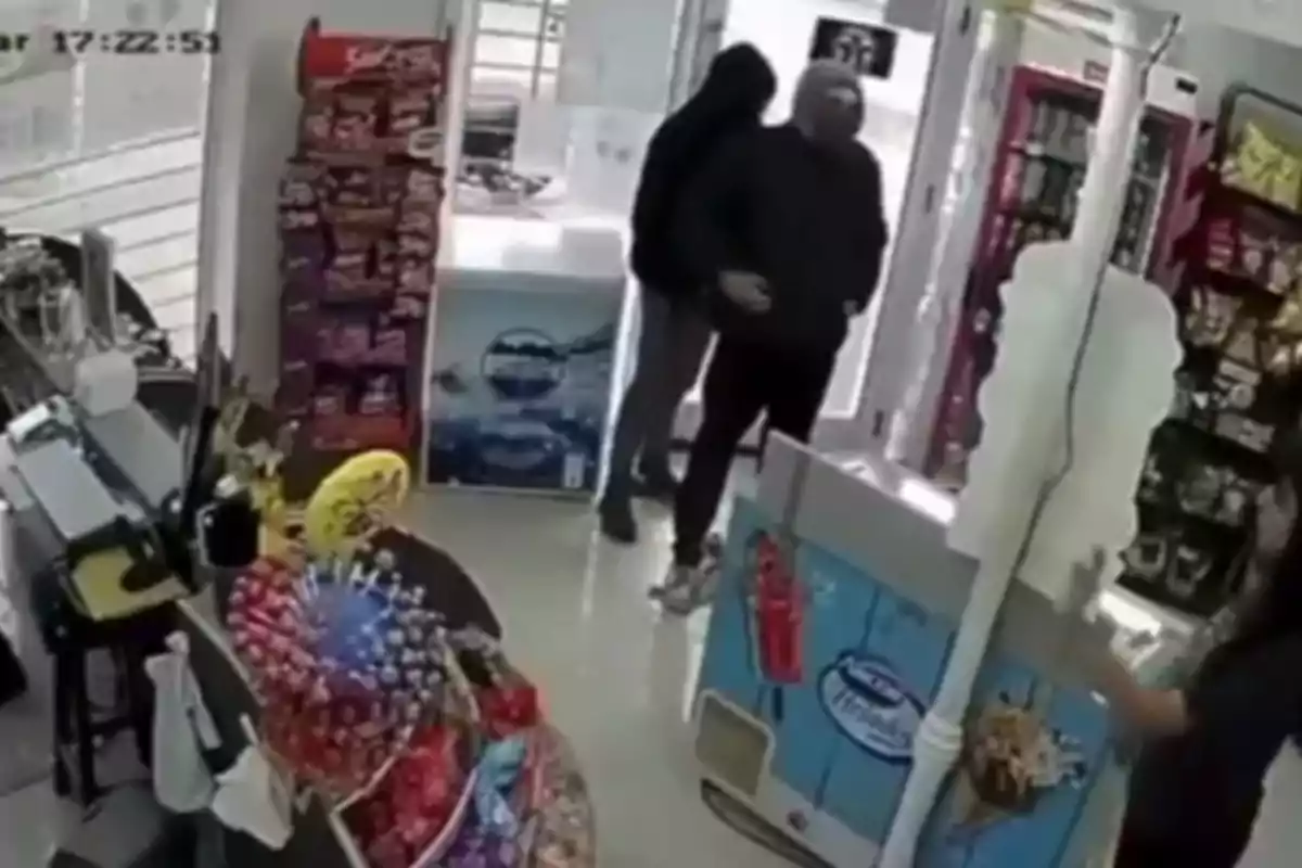 Dos personas con capuchas entran a una tienda mientras una empleada está detrás del mostrador y hay productos en exhibición. Dos personas con capuchas entran a una tienda mientras una empleada está detrás del mostrador y hay productos en exhibición.