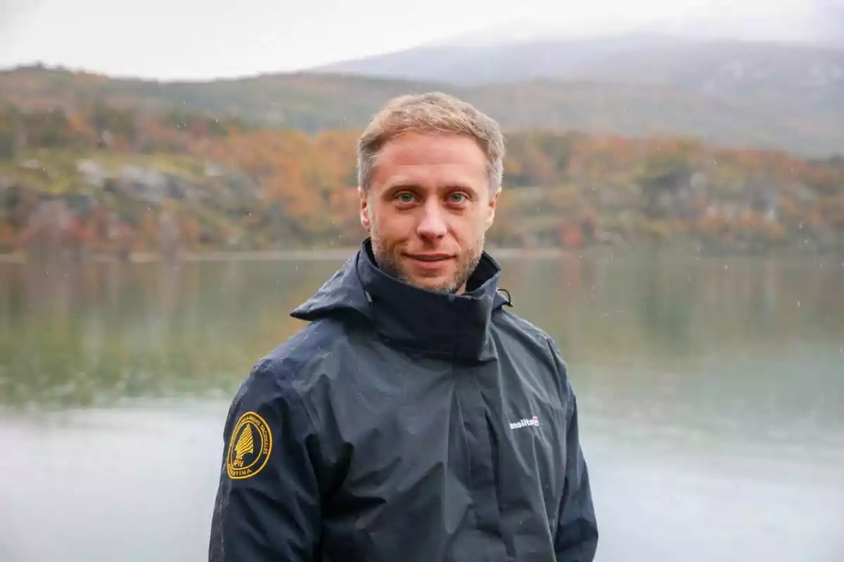 Hombre con chaqueta oscura de pie frente a un lago y montañas con árboles de colores otoñales al fondo Hombre con chaqueta oscura de pie frente a un lago y montañas con árboles de colores otoñales al fondo