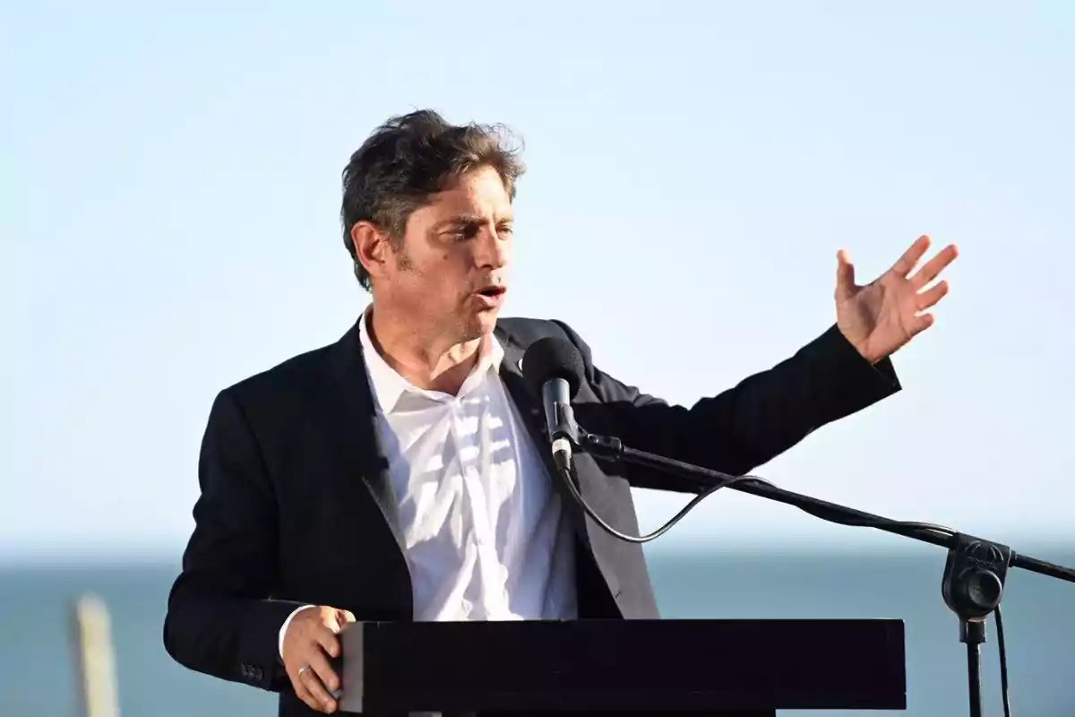 A man in a dark suit speaks into a microphone outdoors with the sea in the background.