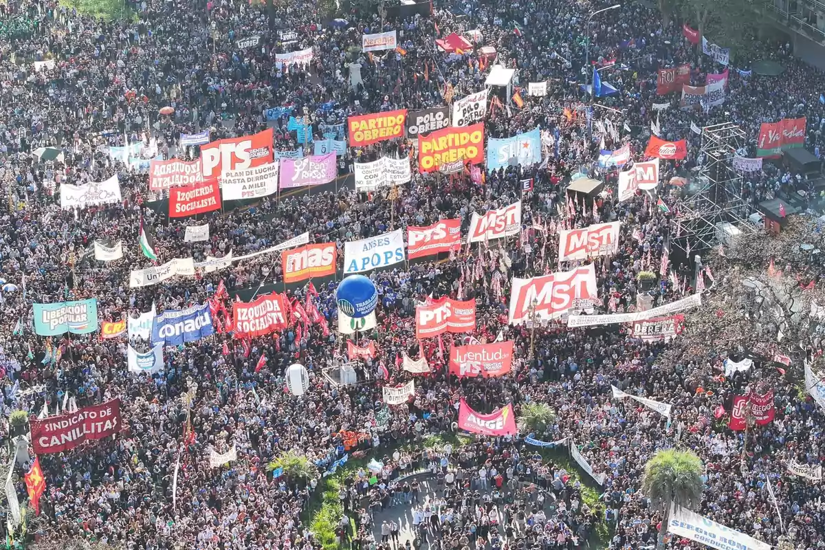 Una multitud de personas se congrega en una manifestación con numerosas pancartas y banderas de diferentes organizaciones y partidos políticos.