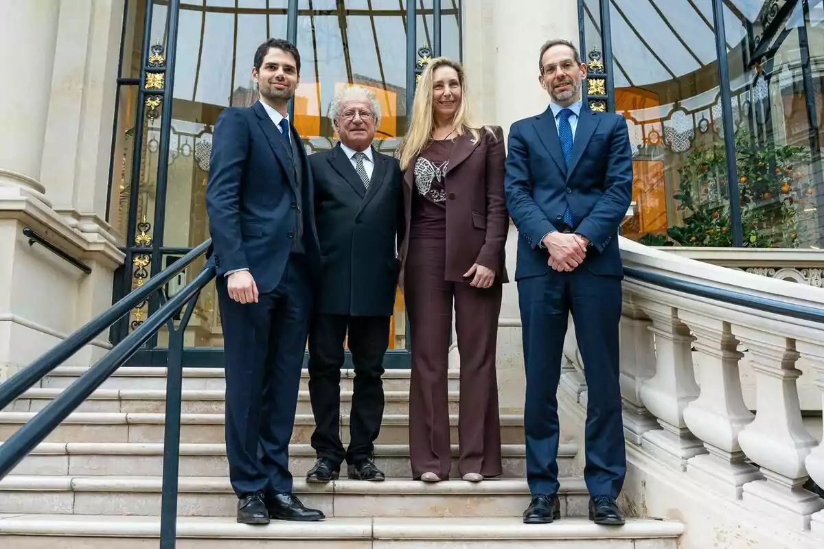 Un grupo de cuatro personas vestidas formalmente posando en una escalera frente a un edificio elegante con una puerta de vidrio y detalles arquitectónicos decorativos.