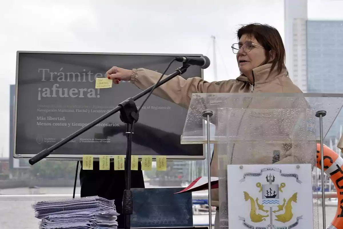 Una persona está de pie junto a un podio transparente con un escudo, señalando una pantalla que dice "Trámite ¡afuera!" y sostiene una nota adhesiva amarilla.