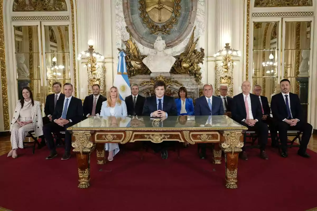 Un grupo de personas vestidas formalmente está sentado alrededor de una mesa en una sala decorada con detalles dorados y una bandera argentina.