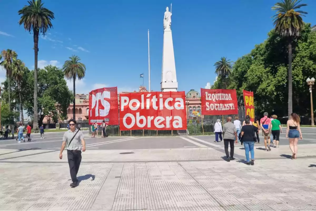 Personas caminando en una plaza con pancartas rojas que dicen 