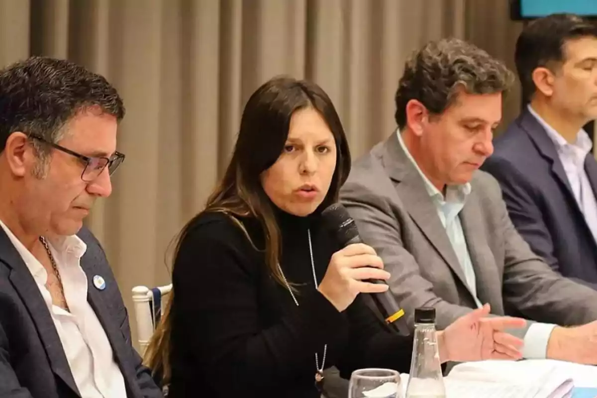 Una mujer hablando por micrófono en una mesa junto a tres hombres en un evento formal.