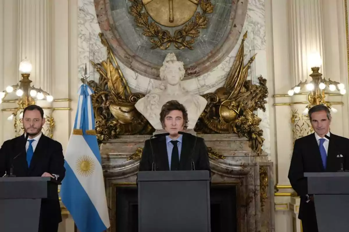 Tres hombres de pie detrás de podios en un salón elegante con una bandera argentina y una escultura decorativa al fondo.