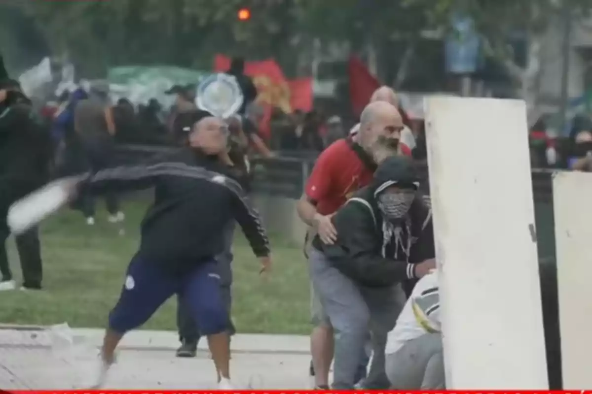 Personas participando en una protesta, algunas con el rostro cubierto, mientras una lanza un objeto y otras se resguardan detrás de una barrera improvisada.