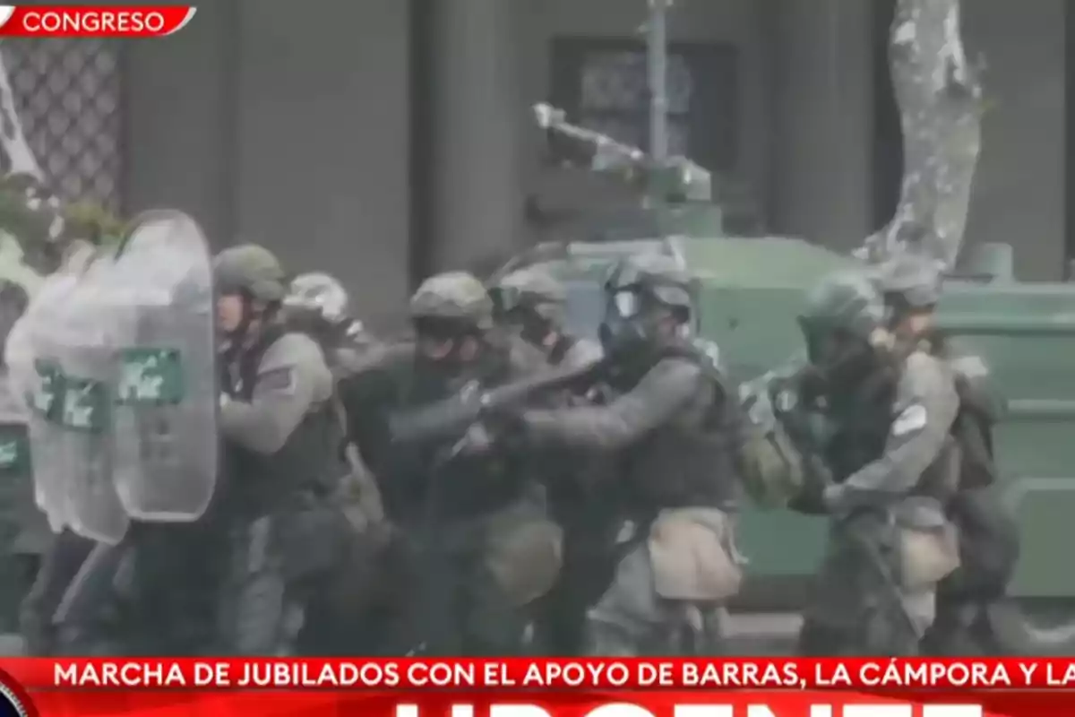 Grupo de policías antidisturbios con equipo de protección avanzando en formación frente a un vehículo blindado.