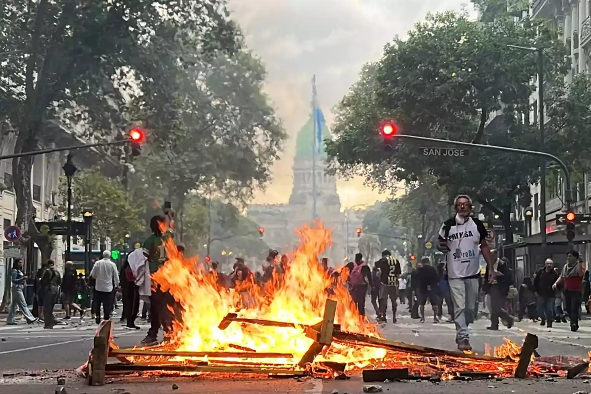 Una protesta en una calle con una barricada en llamas y personas alrededor, con un edificio emblemático al fondo.