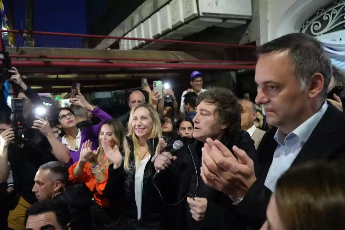 A group of people claps and takes photos while a man holds a microphone at an outdoor event. A group of people claps and takes photos while a man holds a microphone at an outdoor event.