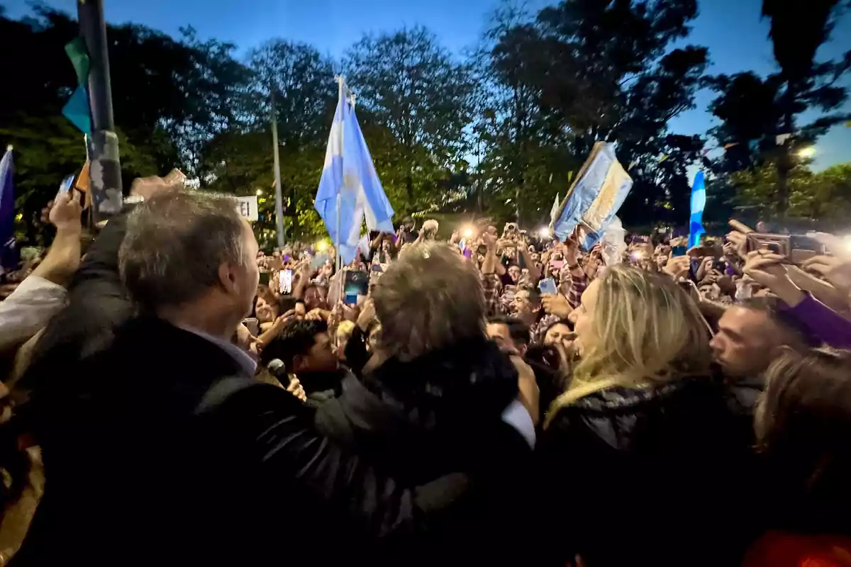 A crowd of people gathered outdoors holding Argentine flags and taking photos with their phones. A crowd of people gathered outdoors holding Argentine flags and taking photos with their phones.