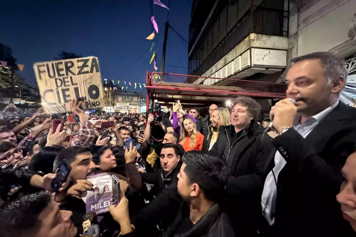 A crowd gathers at an outdoor event while a person speaks into a microphone and others hold signs and mobile phones.