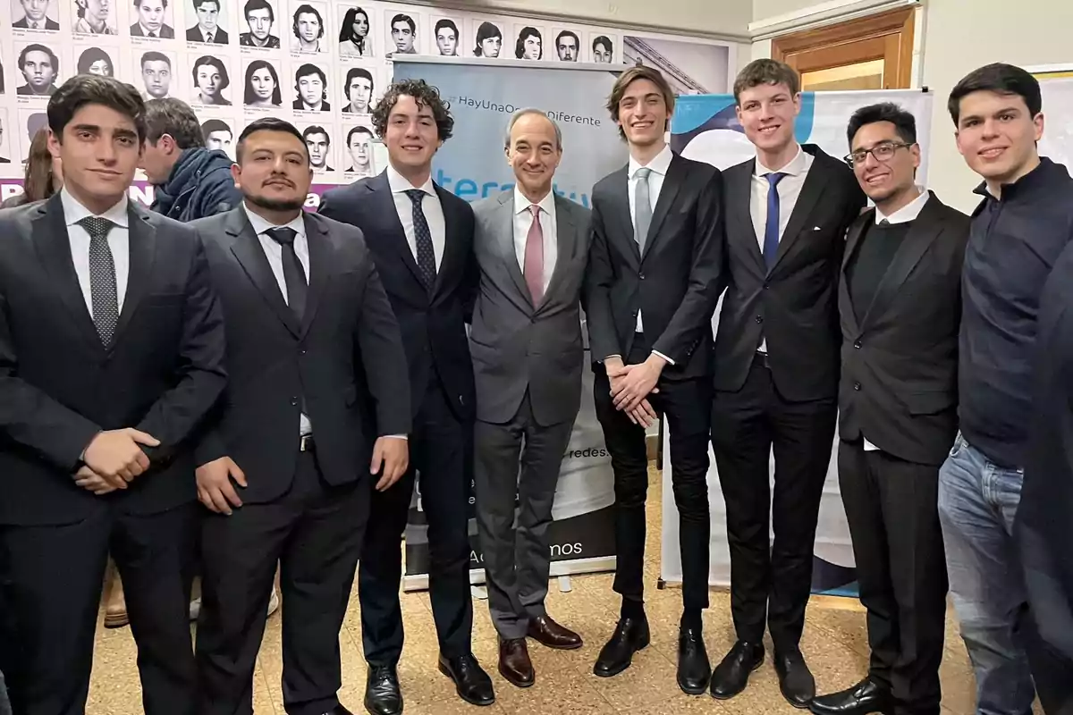 Un grupo de hombres vestidos de traje posan juntos para una foto en un evento formal con retratos en blanco y negro en la pared de fondo. Un grupo de hombres vestidos de traje posan juntos para una foto en un evento formal con retratos en blanco y negro en la pared de fondo.
