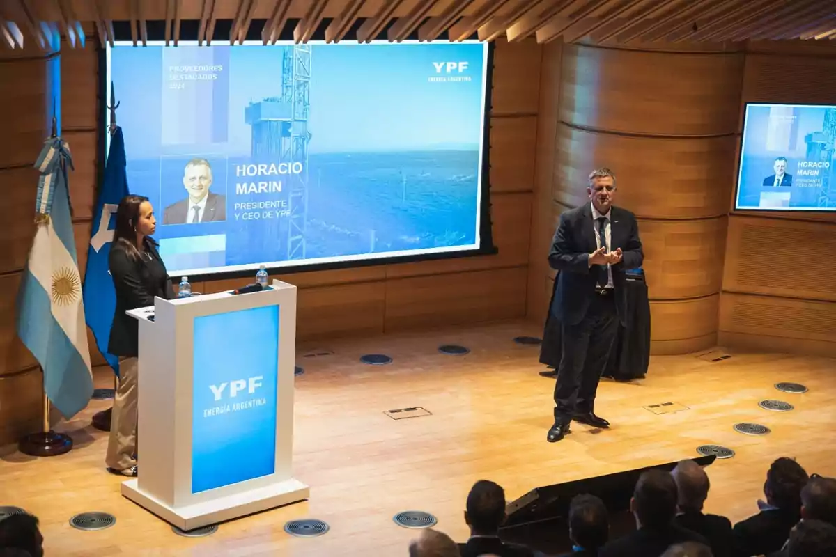 A corporate presentation in an auditorium with a man speaking and a woman at a podium, with screens displaying information about YPF and Argentine flags. A corporate presentation in an auditorium with a man speaking and a woman at a podium, with screens displaying information about YPF and Argentine flags.