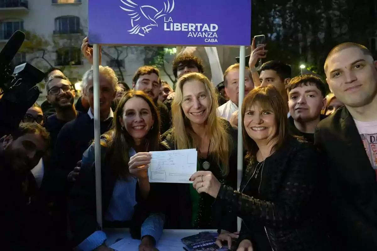 Un grupo de personas sonríe mientras sostiene un documento frente a un cartel que dice "La Libertad Avanza CABA" en un evento al aire libre.